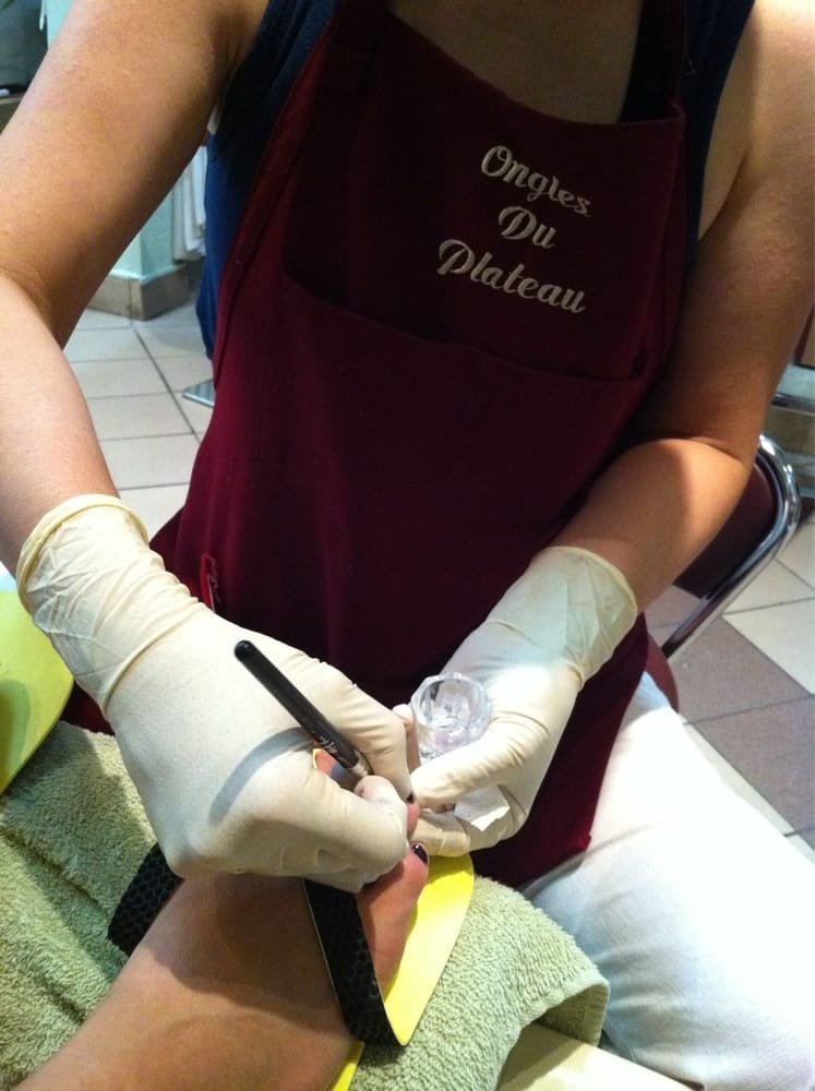 Technicienne en tablier brodé Ongles Du Plateau réalisant une pose d'ongles gel avec des gants.